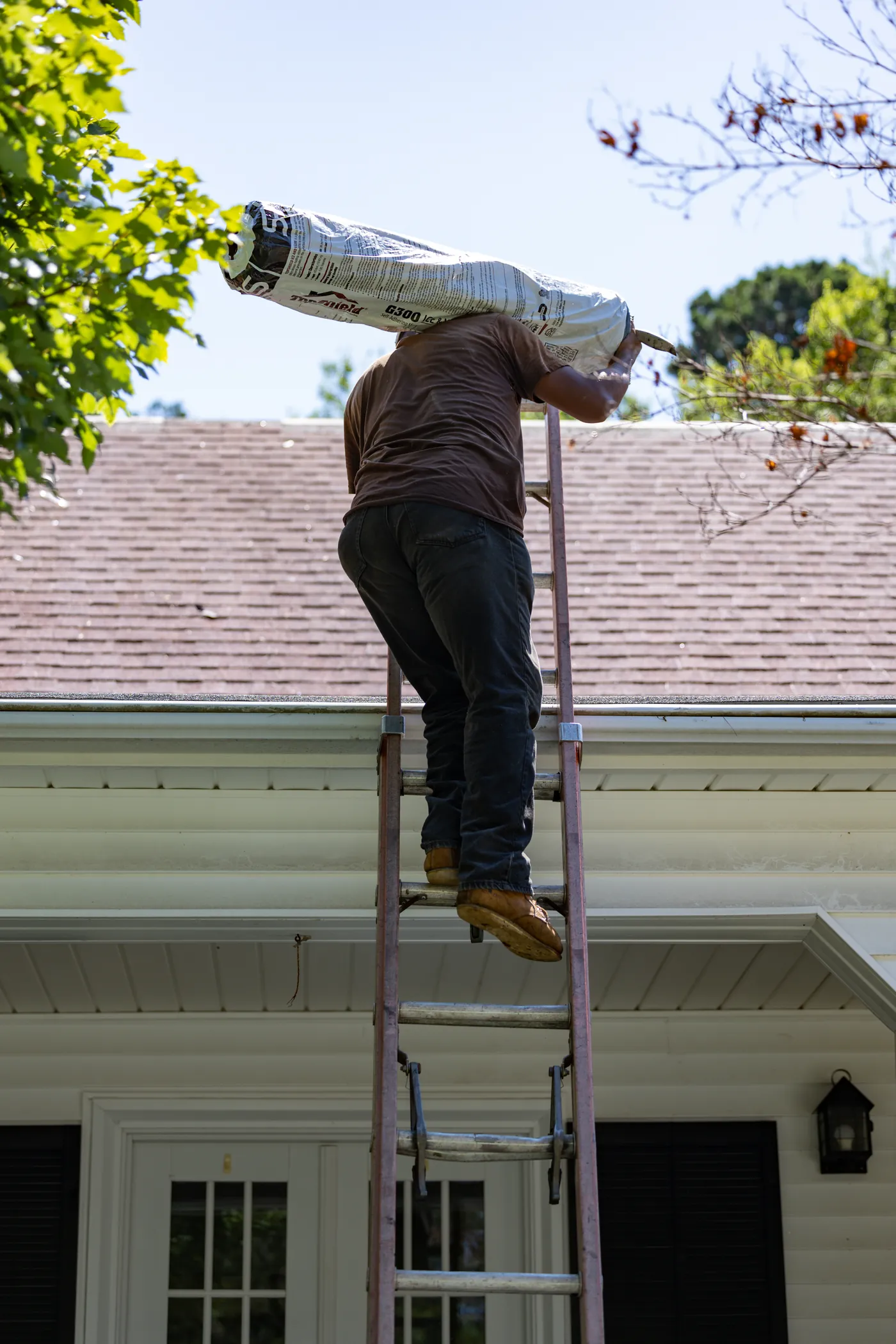 Brown's Roofing crew member climbing a ladder with a shingle bundle on his shoulder on a Bryant area job site